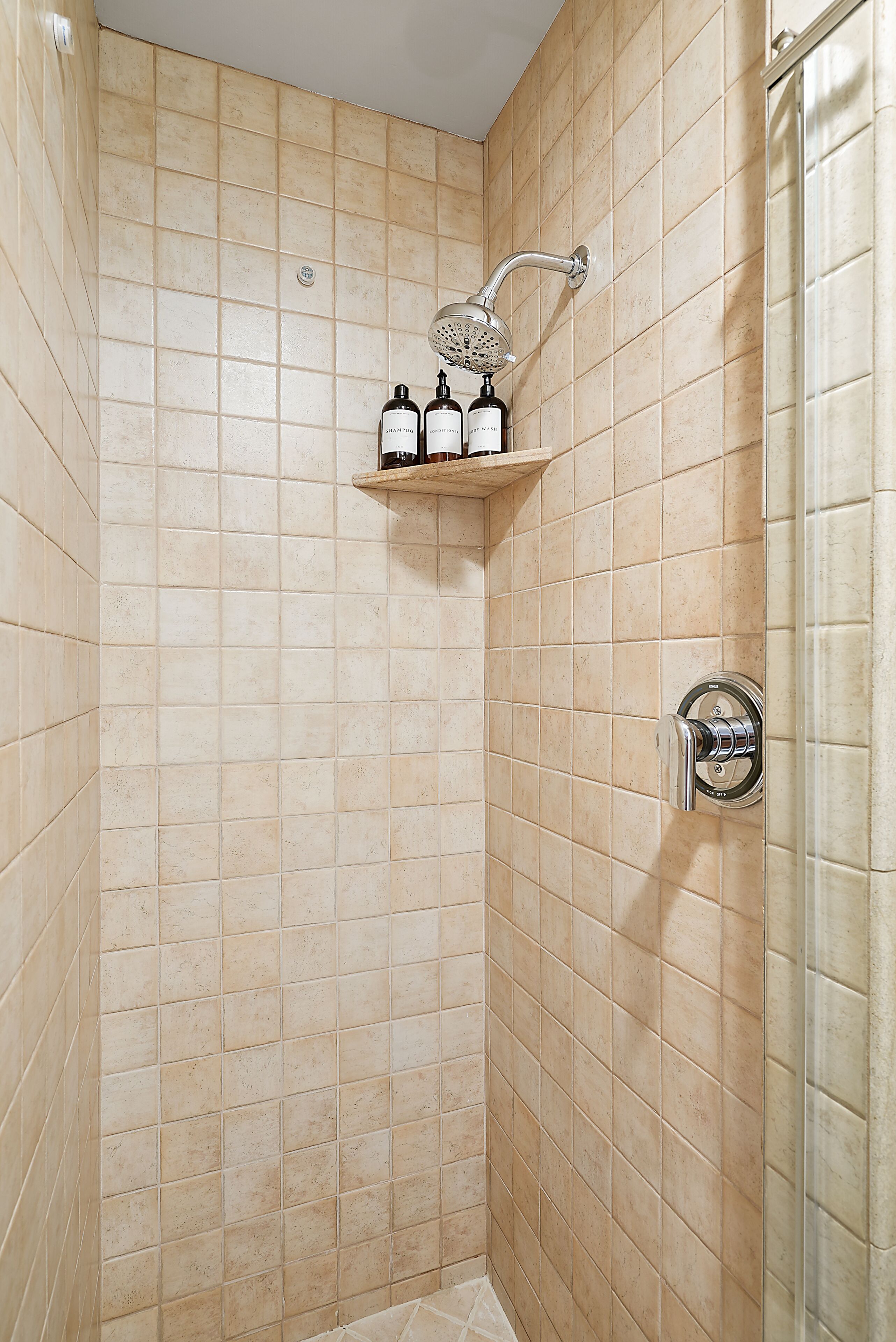 Una ducha con azulejos color crema, un cabezal de ducha manual y un estante con artículos de aseo.