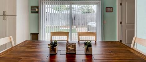 A dining room with a wooden table, two chairs, two hanging pendant lights, and a view through a sliding glass door to a backyard.
