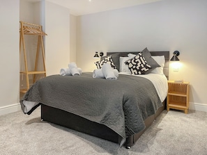 Una cama bien tendida con un edredón oscuro, una escalera de madera y una mesita de noche con una lámpara.