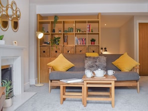 Una sala de estar acogedora con un sofá gris, cojines amarillos, una mesa de centro de madera y una estantería llena de libros y objetos decorativos.