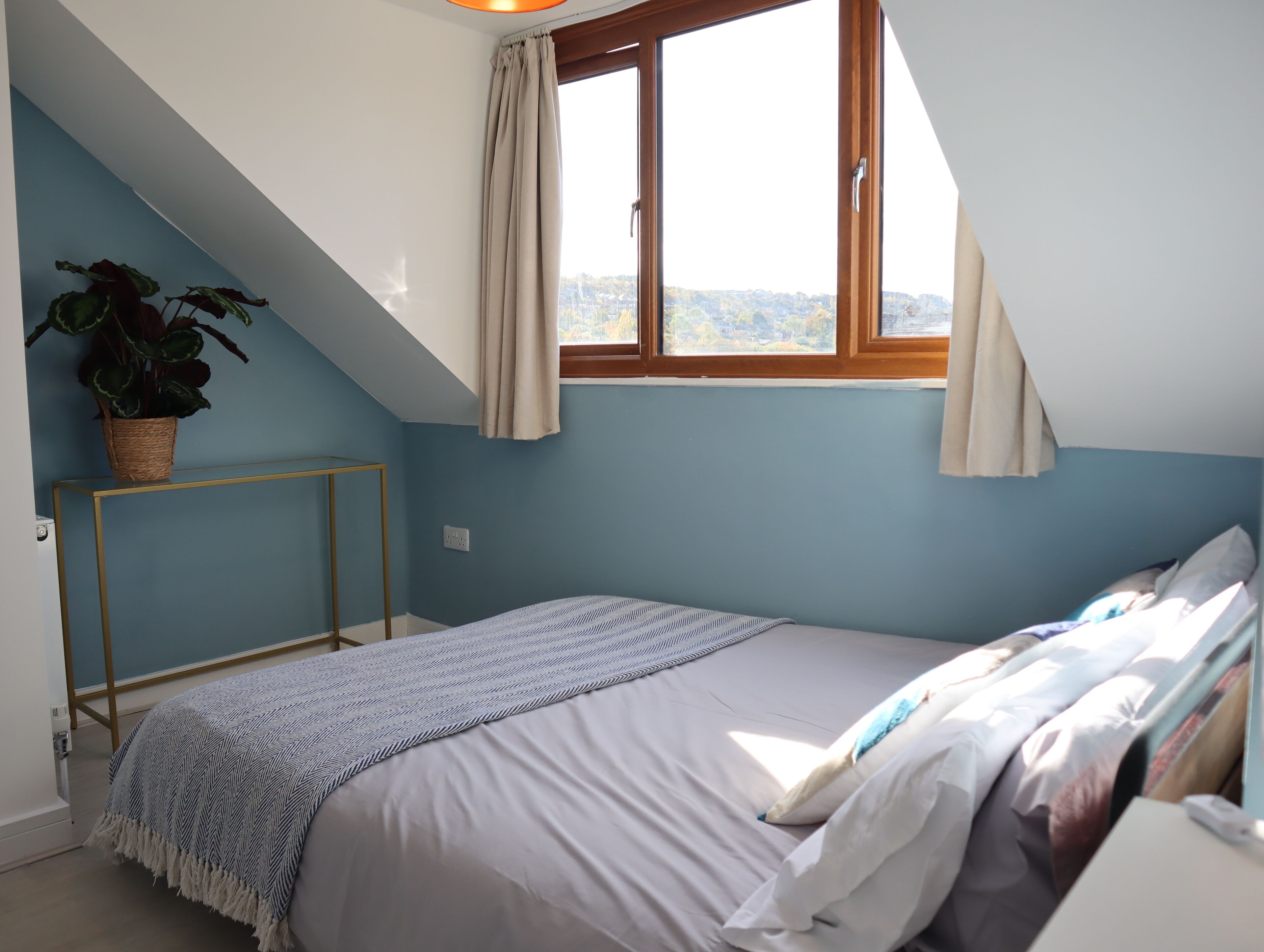 Un dormitorio con techo inclinado, una cama con manta estampada, una planta en maceta y una ventana con cortinas.