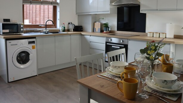 Une cuisine moderne équipée d’un lave-linge, d’un four à micro-ondes et d’un four traditionnel. On y trouve également un coin repas avec une table dressée pour deux personnes, comprenant des tasses jaunes et un vase avec des fleurs.