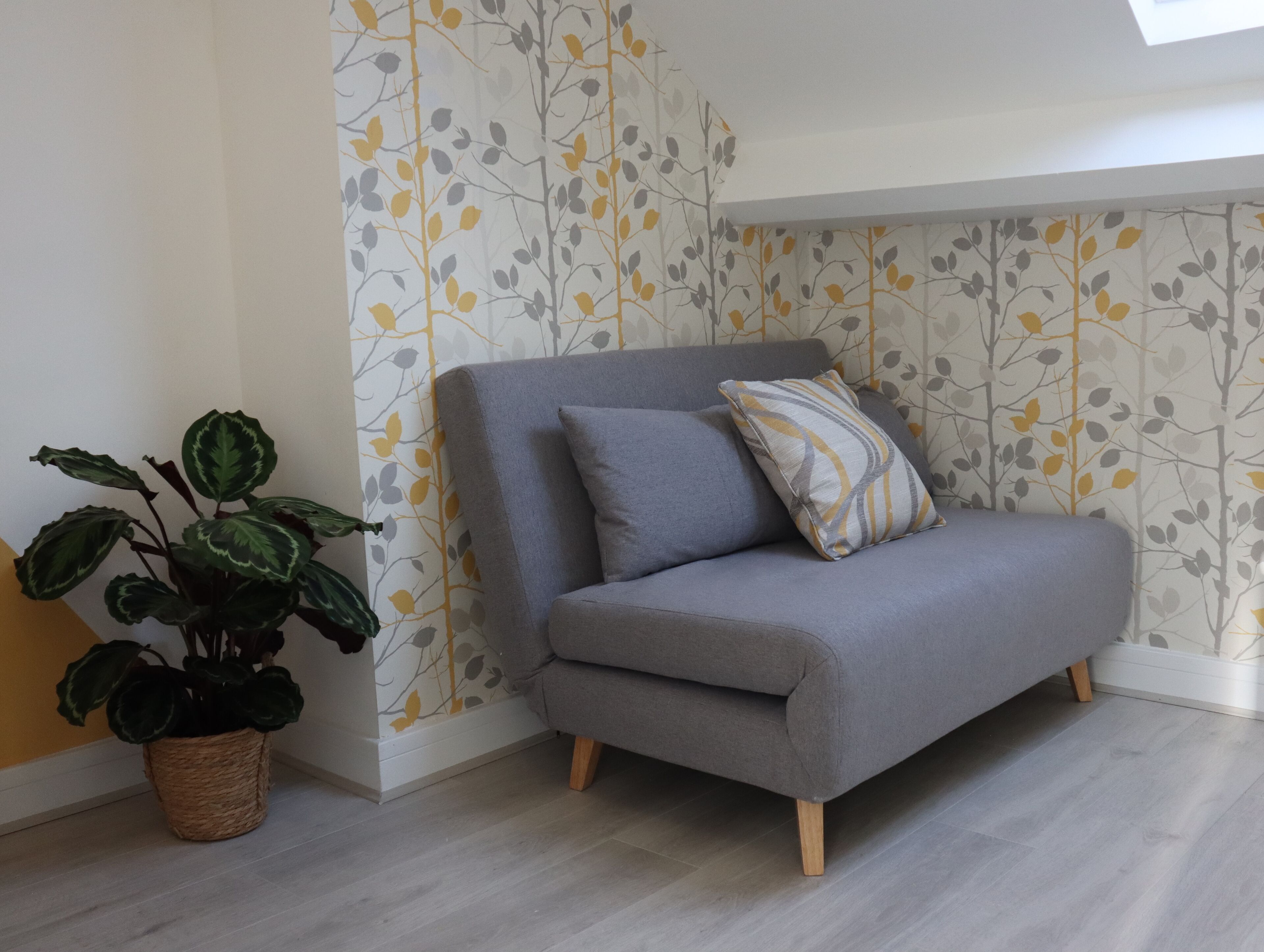 Ein graues Sofa mit Holzbeinen, ein Blumentopf und eine Wand mit einem Blattmuster.