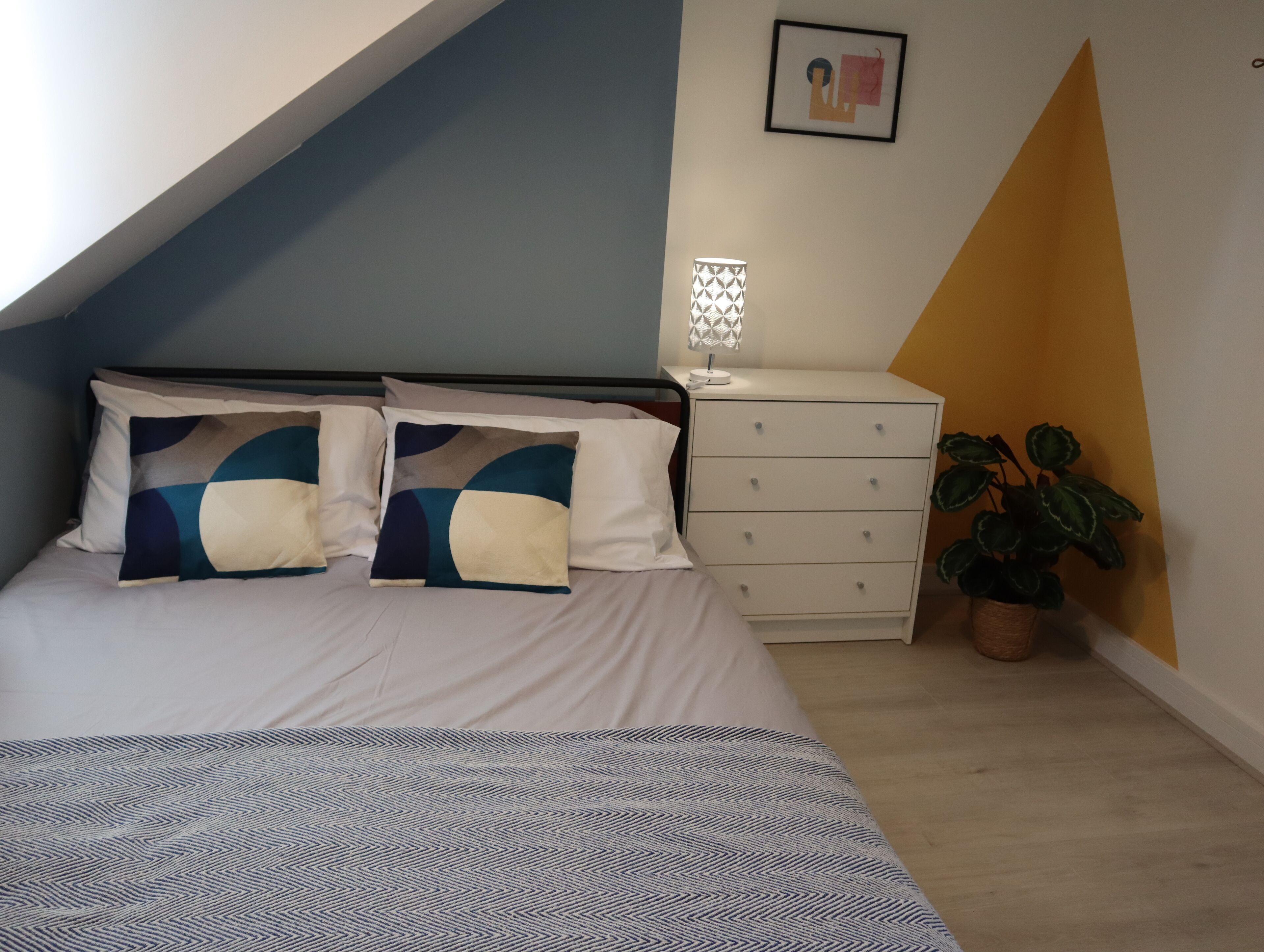 A bedroom with a bed, a bedside table, a lamp, and a potted plant.