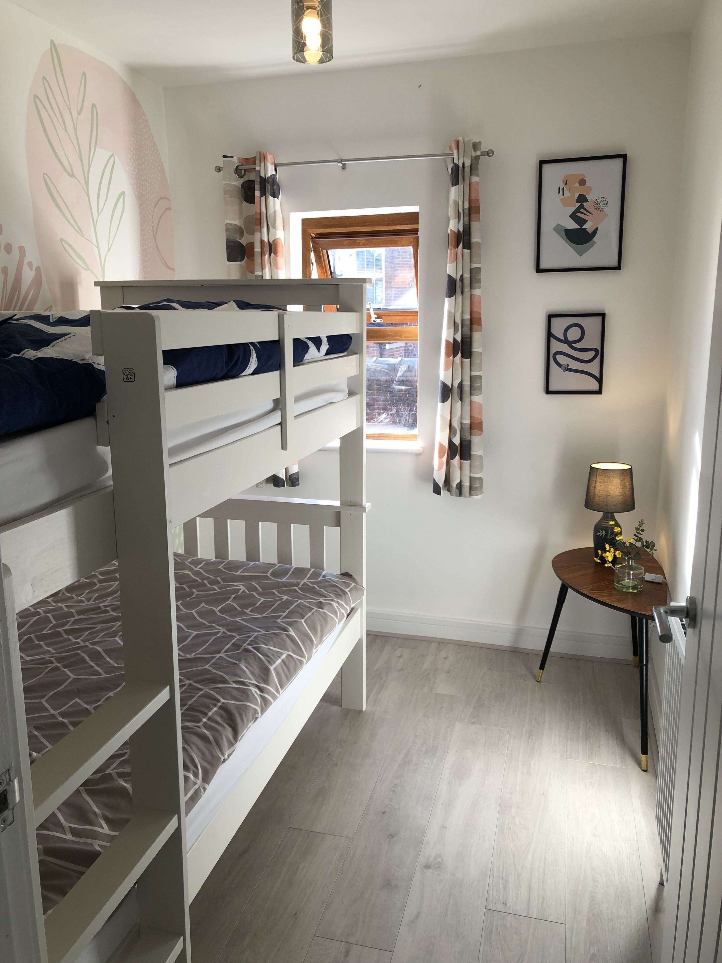 A bunk bed room with a window, a small table with a lamp, and wall art.
