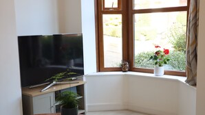 Une chambre d’angle avec un téléviseur posé sur un meuble, une petite table avec une plante et une fenêtre donnant sur un paysage verdoyant.