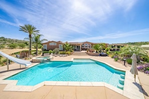 A swimming pool with a slide, surrounded by a landscaped garden and a building with a patio.