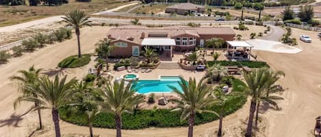 A large house with a swimming pool, surrounded by palm trees and a dirt road.
