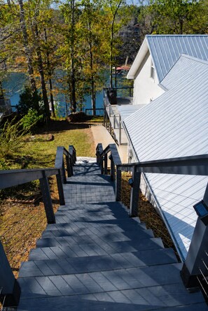 A staircase leading to a building with a metal roof, surrounded by trees and a body of water. - The Gathering Place: Big Lakehouse/Covered Dock/Fiber/Tankless WH/Game RM/EV CHG (Cullman)