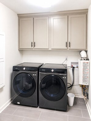 Une buanderie équipée de deux machines à laver Samsung noires, de meubles de couleur beige et d’un sol carrelé blanc.