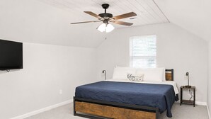 Un dormitorio con cama, ventilador de techo, televisor montado en la pared y ventana con persianas.