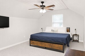 Un dormitorio con cama, ventilador de techo, televisor montado en la pared y ventana con persianas.
