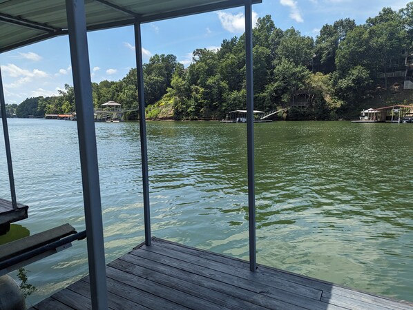 Un muelle de madera se extiende sobre un lago tranquilo, con una barandilla de metal. Al fondo, se ven edificios con techo rojo y un paseo cubierto.