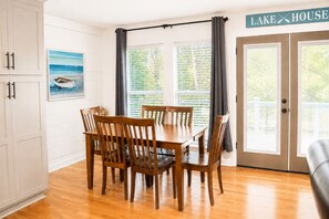 Un coin repas avec une table et des chaises en bois, un tableau représentant un bateau sur l’eau et une pancarte indiquant « MAISON AU BORD DU LAC ».