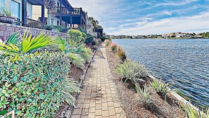 En havnefrontpromenade med belagt sti, omkranset af frodig vegetation og palmer, der fører til en moderne bygning med balkoner med udsigt over vandet.