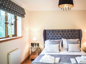 Ett sovrum med en stoppad sänggavel, ett sängbord med en lampa, ett element och ett fönster med utsikt över träd.