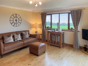 A living room with a brown leather sofa, a wooden coffee table, a clock, and a view of a green field through a window.