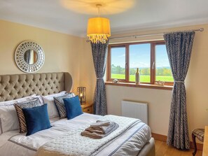 Ein Schlafzimmer mit einer gesteppten Kopfteil, einem Bett mit weißer Bettwäsche und blauen Kissen, einem Nachttisch mit Lampe, einem Fenster mit Vorhängen und Blick auf Grünfläche außerhalb.