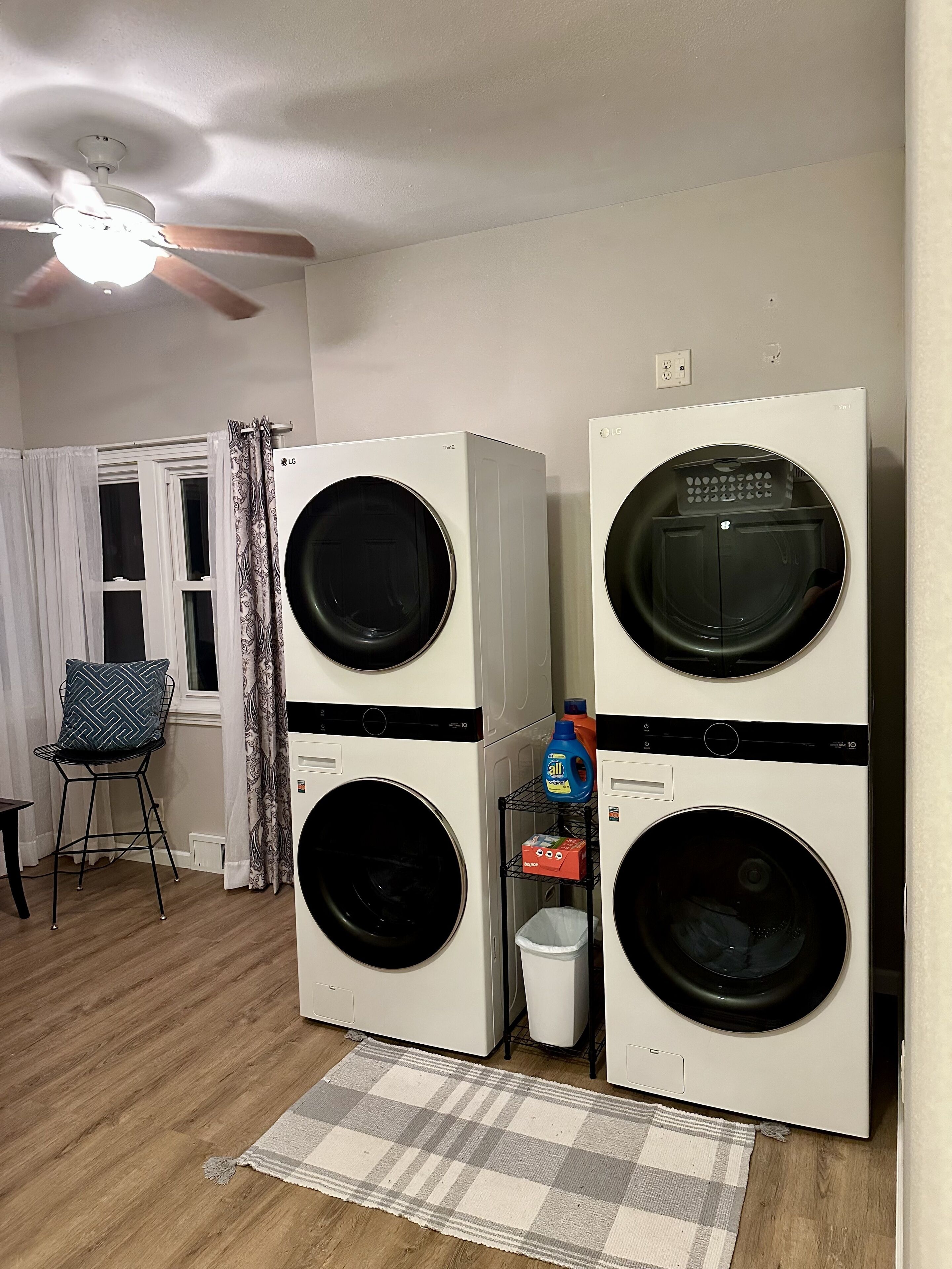 A laundry room with stacked front-loading washers and dryers, a ceiling fan, a window with curtains, and a small chair.