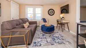 Un salon comprenant un canapé, un pouf et une table basse, un coin repas avec une table et des chaises, et une horloge fixée au mur.