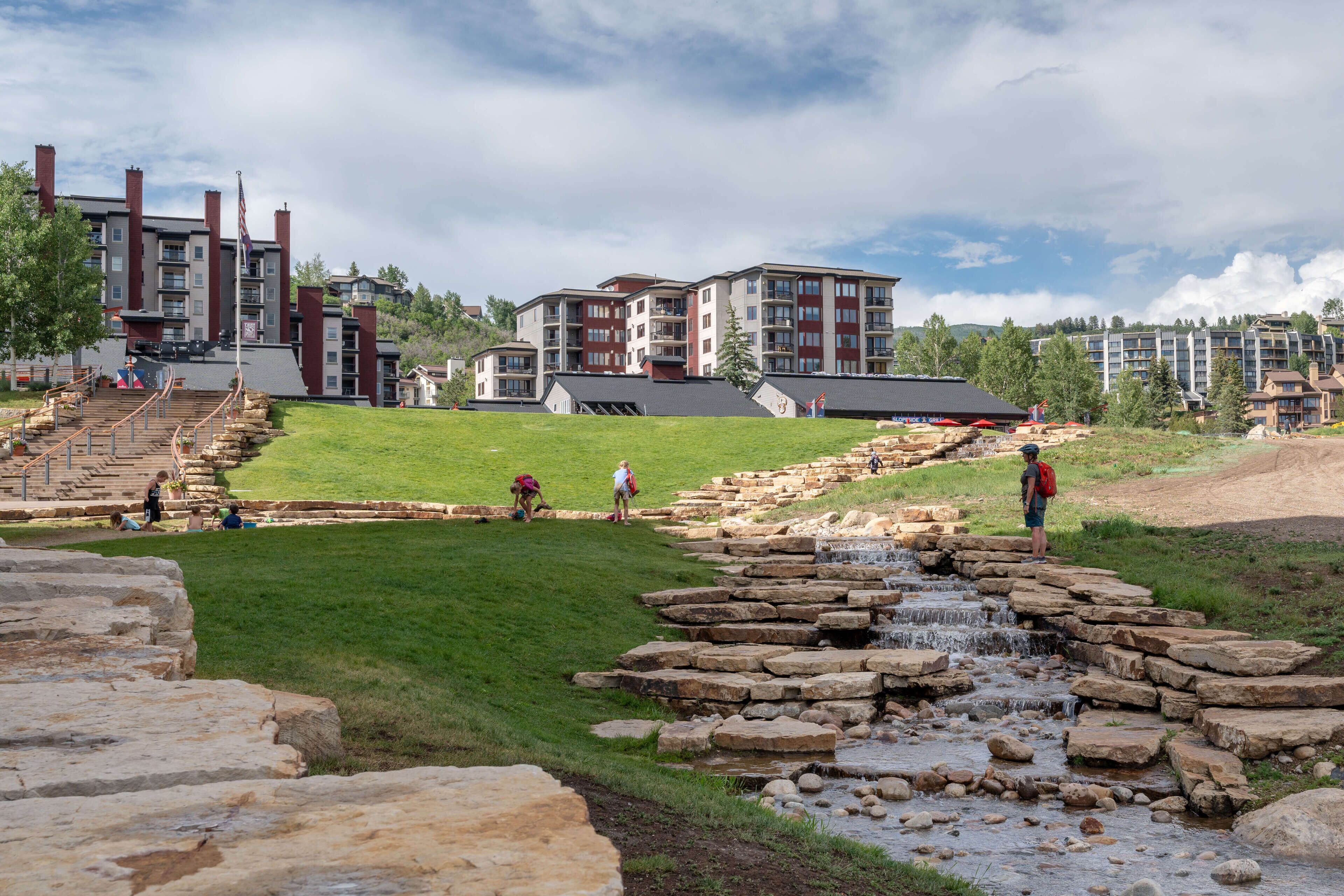 A modern hotel with a stone pathway leading to a small waterfall, surrounded by lush greenery and a clear sky.