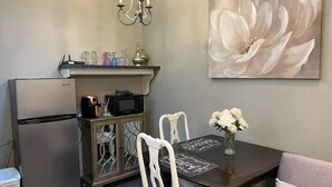 Une cuisine compacte équipée d’un réfrigérateur, d’un four à micro-ondes et d’un meuble avec des portes en verre. On y trouve une table à manger avec des chaises et un vase de fleurs. Un lustre est suspendu au-dessus de la cuisine, et un grand tableau représentant des fleurs orne le mur.