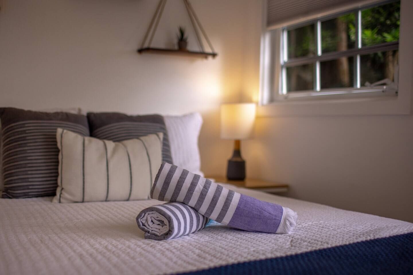 A neatly made bed with striped and solid pillows, a rolled-up towel, and a bedside lamp.
