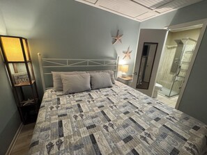 A bedroom with a bed, a nightstand, a lamp, a starfish decoration, and a shower area. - Paradise PRN (gulf shores)