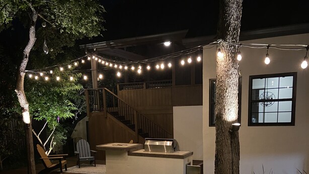 Un espace patio agrémenté de guirlandes lumineuses, une terrasse en bois, un chemin de pierre et un petit coin salon avec des chaises.