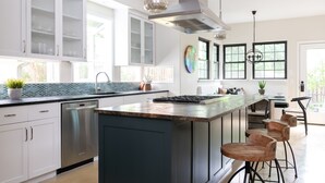 Une cuisine moderne avec des armoires blanches, un lave-vaisselle en acier inoxydable, un grand îlot central doté de tabourets de bar et une vue sur l’extérieur grâce à de grandes fenêtres.