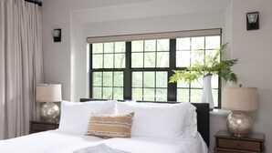 Une chambre avec un grand lit, deux tables de chevet avec des lampes, une fenêtre donnant sur un beau paysage, un ventilateur de plafond et un pouf de couleur sombre.