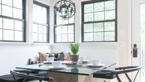 Une cuisine moderne avec une table à plateau en verre, deux chaises, un luminaire suspendu et de grandes fenêtres.