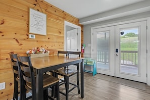 A dining area with a wooden wall, a framed quote, a table with chairs, and a view of a green outdoor area through a door. - Ridge top cabin with hot tub and movie theater (Logan)