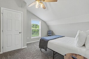 A bedroom with a bed, a ceiling fan, a window with blinds, a small table, and a remote control.