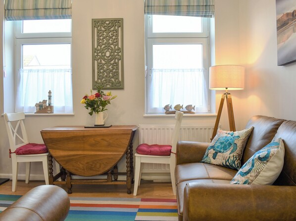 A cozy living room with a brown leather sofa, a wooden dining table, and a white chair. There is a lamp on a floor lamp stand, a colorful striped rug, and a decorative wall piece above the windows.