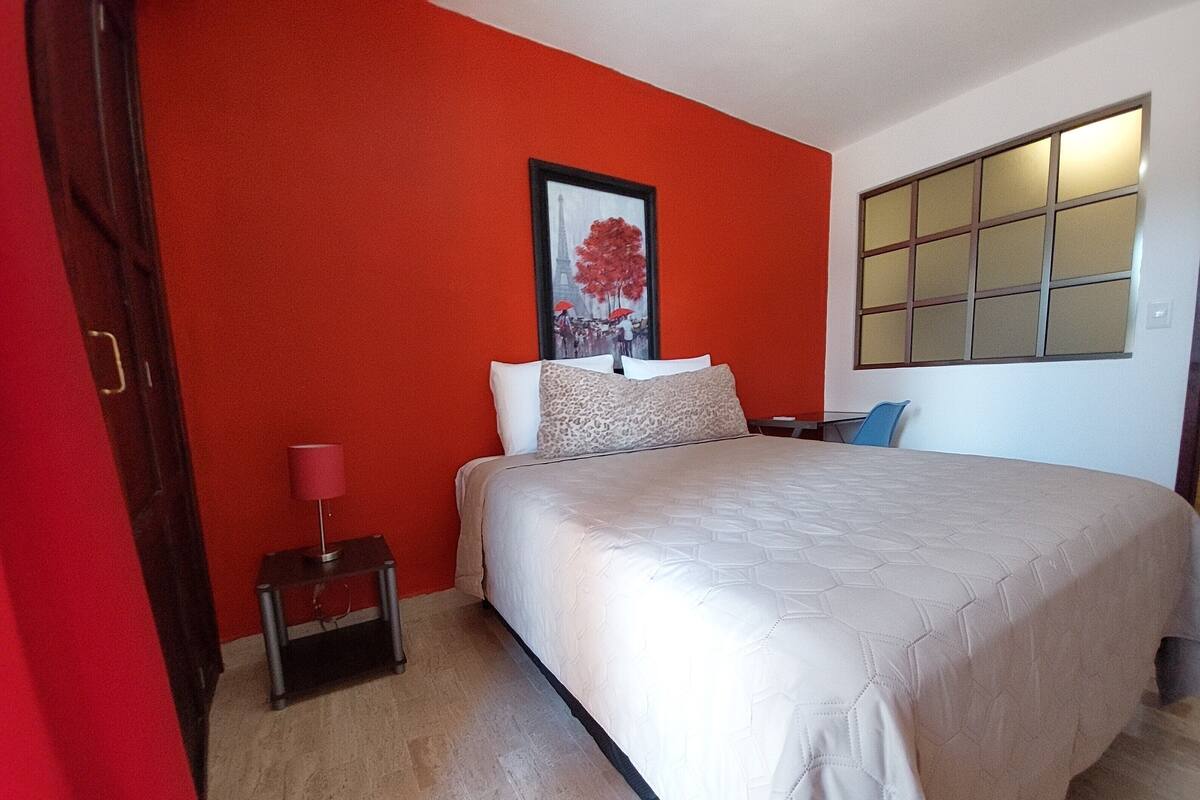 A bedroom with a large bed, a red wall, a framed picture of the Eiffel Tower, a nightstand with a lamp, and a window with blinds.