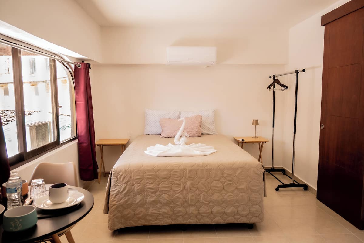 A hotel room with a bed, a table with a cup and saucer, a chair, a window with curtains, an air conditioning unit, and a floor lamp.