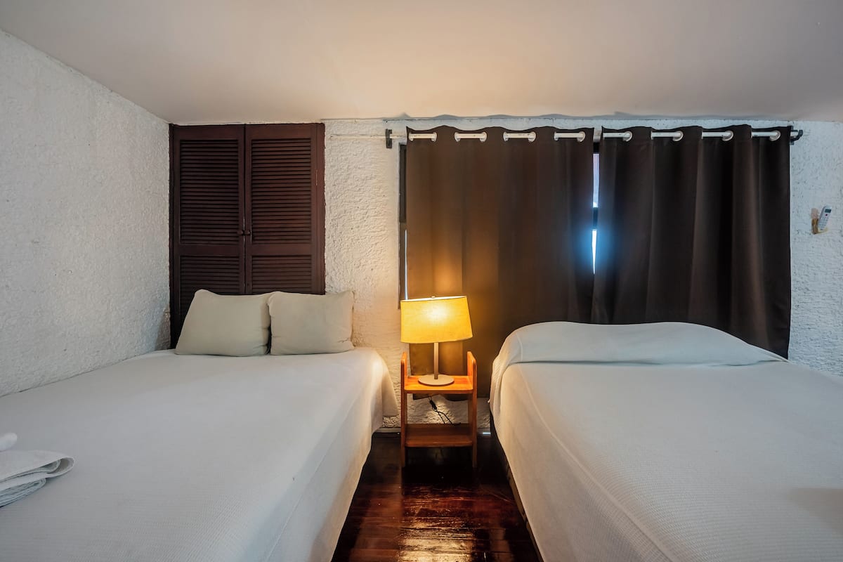 A hotel room with two beds, a nightstand with a lamp, wooden shutters, and a curtain.