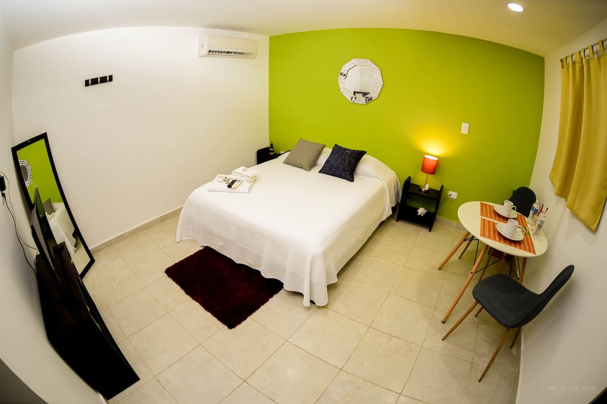 A hotel room with a bed, bedside table, chair, and a small table with a lamp.