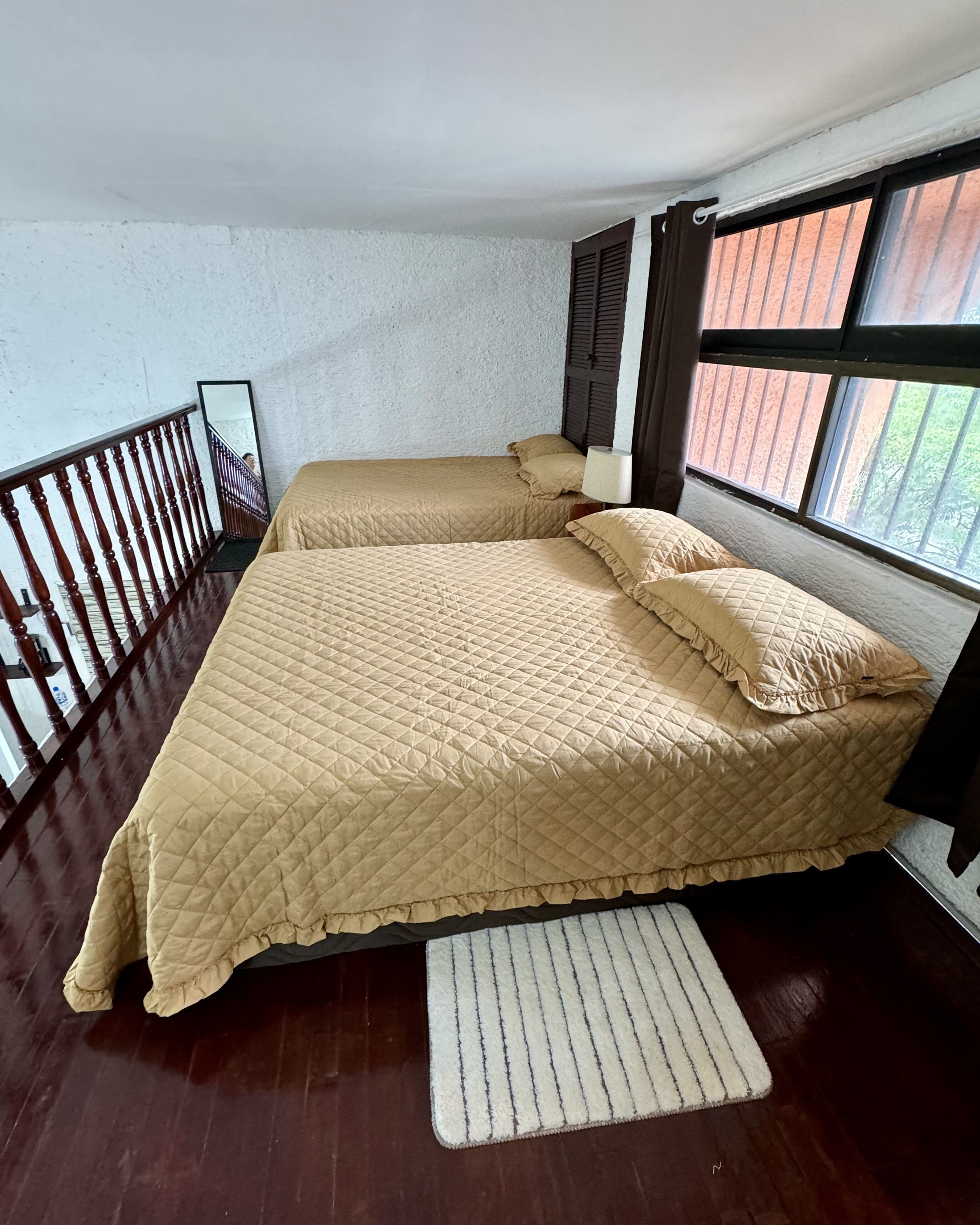 A room with two beds, a wooden floor, a white wall, and a window with blinds.