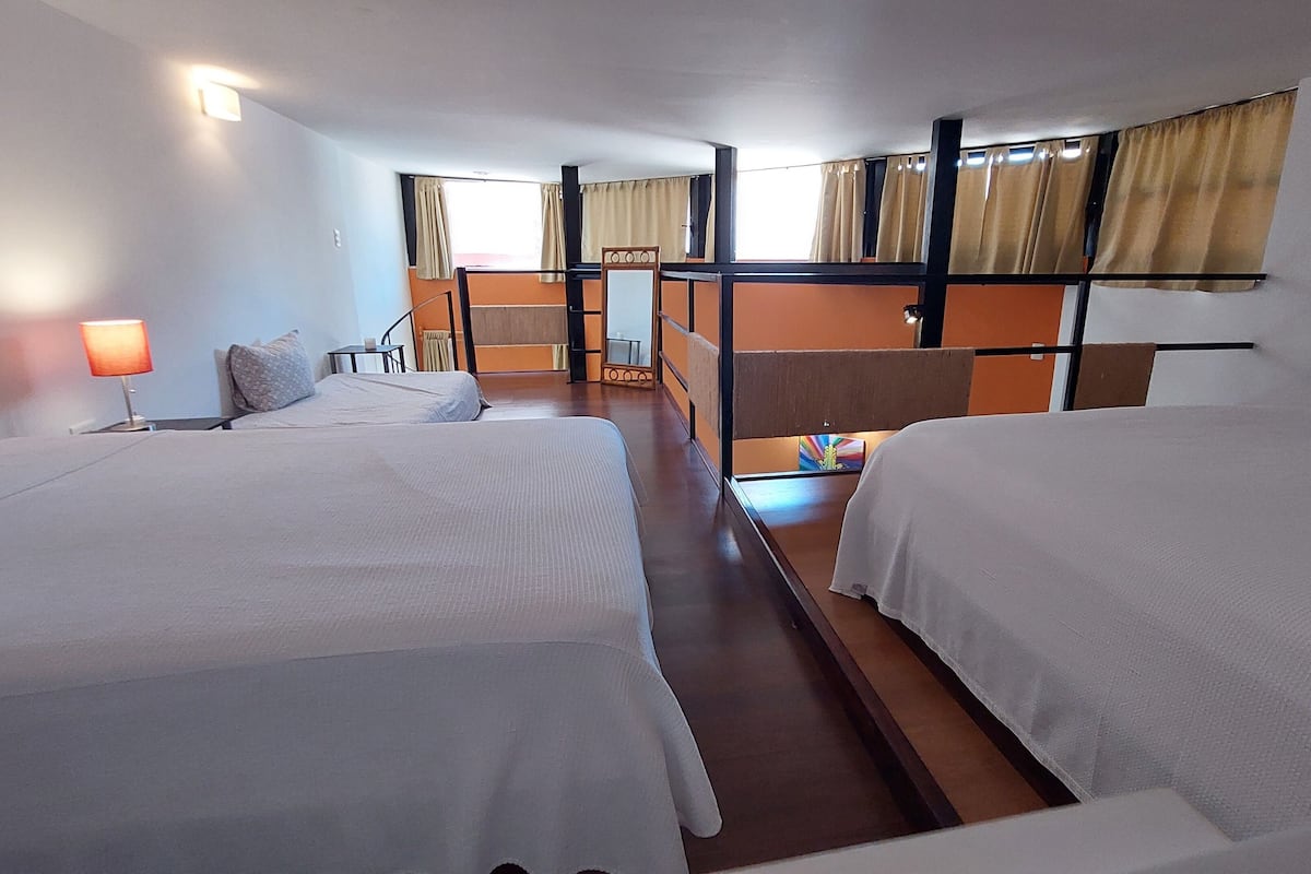 A hotel room with two beds, a small table with a lamp, and a window with curtains.
