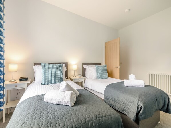 A hotel room with two beds, each with a pillow and a blanket. There are bedside tables with lamps and a clock.