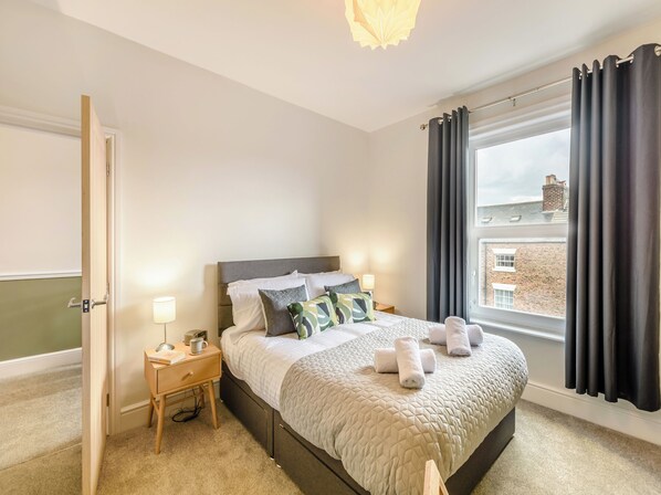 A neatly arranged bedroom with a bed, bedside table, lamp, and a view of the outside.