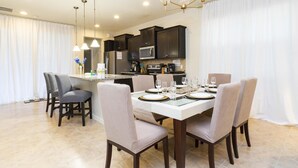 Un espace repas moderne avec une table blanche dressée pour six personnes, entourée de chaises rembourrées. On aperçoit en arrière-plan une cuisine avec des meubles sombres et des appareils en acier inoxydable.
