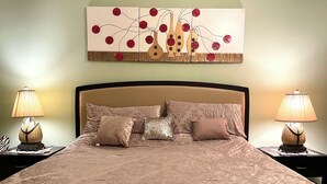 Uma cama bem arrumada com colcha estampada, ladeada por duas mesas de cabeceira com abajures, e um grande quadro abstrato acima da cama.