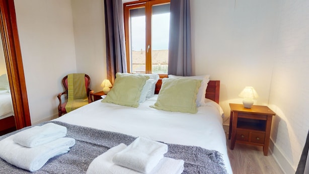 Hotellihuone, jossa on sänky, kaksi tyynyä, tuoli, yöpöytä lampun kanssa ja ikkuna, jossa on verhot.