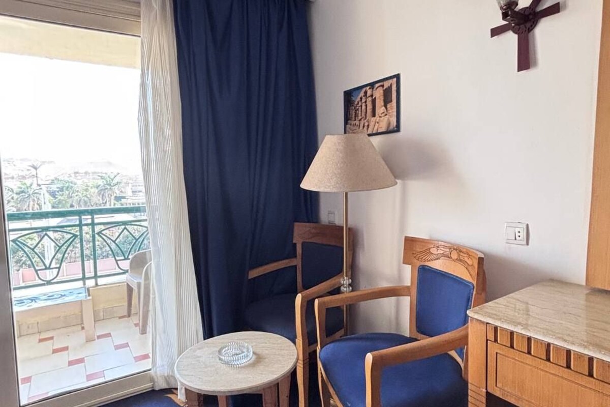 A hotel room with a balcony, a round table, two chairs, a desk, a lamp, and a wall-mounted decoration.