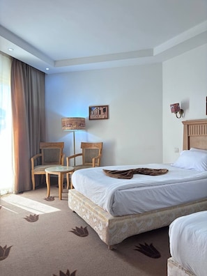 A hotel room with a bed, two chairs, a small table, a lamp, and a wall-mounted light fixture. - A pyramids view (Giza)