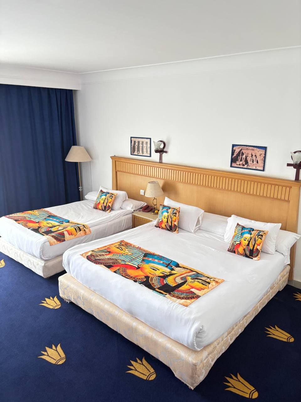 Une chambre d’hôtel avec deux lits simples, une tête de lit en bois, un rideau bleu, une lampe et deux tableaux encadrés accrochés au mur.