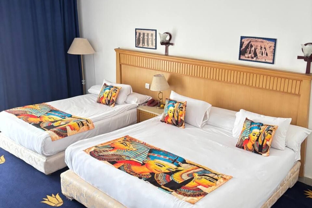 A hotel room with two single beds, a wooden headboard, a blue curtain, a lamp, and two framed pictures on the wall.